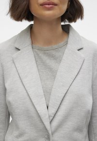 Světle šedý blazer s klopami, texturovanou tkaninou a zapínáním na jeden knoflík. Nosit přes světle šedé žebrované tričko.