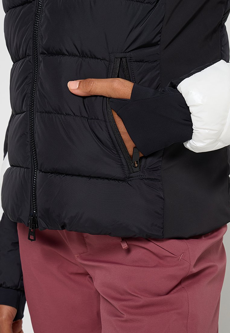 Schwarze Pufferjacke mit horizontaler Steppung, Reißverschlusstaschen und weißen Akzenten an den Ärmeln. Weicher Stoff und elastische Bündchen. Getragen über rosa Hosen.