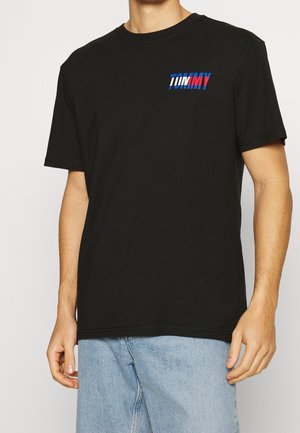 Homme portant un t-shirt noir à manches courtes avec "TOMMY" en lettres bleues et rouges en gras sur le côté gauche de la poitrine, assorti à un jean bleu clair.