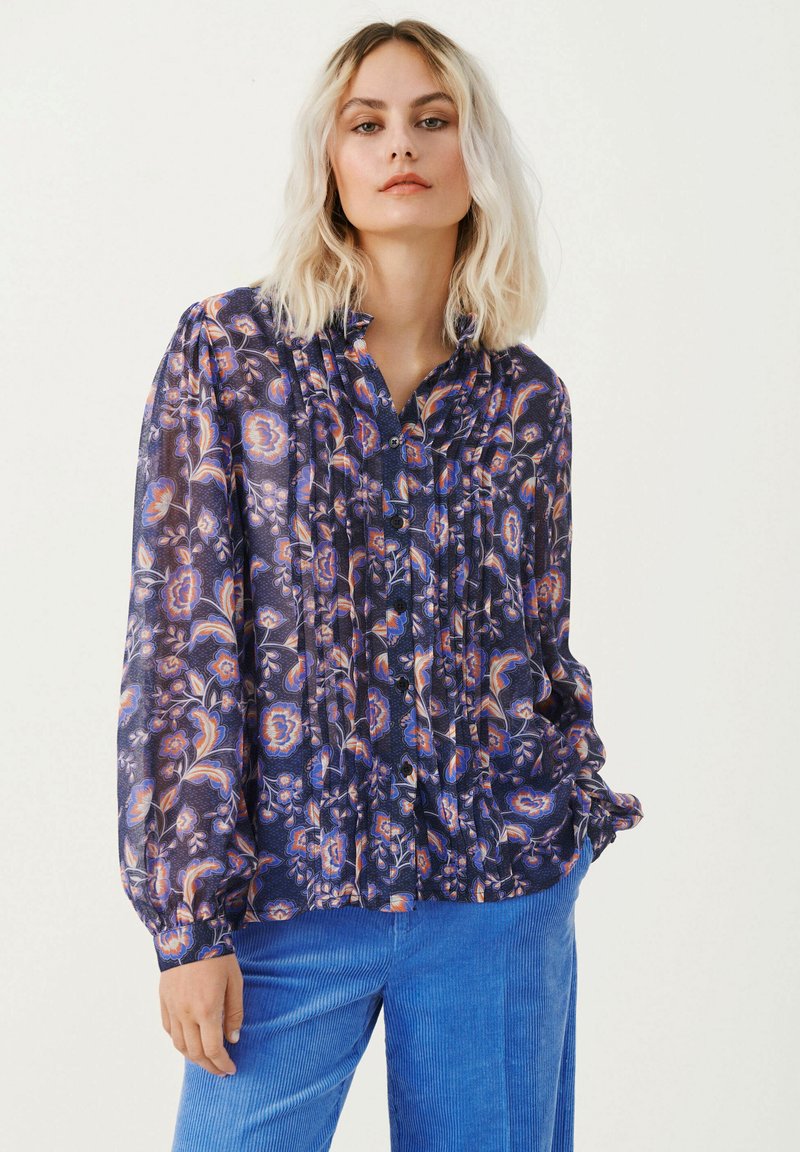 Kobieta z włosami blondu do ramion, ubrana w ciemną, kwiecistą bluzkę z guzikami i jasnoniebieskie spodnie z korduroju, stojąca na tle jednolitym.