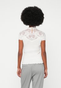 Top blanco de canalé con yugo de encaje y mangas cortas, que presenta un detalle de abertura en la espalda. El dobladillo con borde en picos añade textura. Combinado con pantalones de rayas grises.