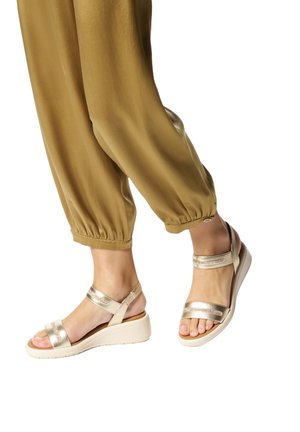 Piedi indossando sandali con zeppa beige e cinturini alla caviglia, abbinati a pantaloni larghi color senape che finiscono sopra le caviglie.
