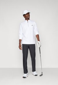 Biela dlhá rukávová košeľa s štvrťzipom a sivým logom, tmavosivé nohavice, biela čiapka, biele golfové topánky a drží golfovú palicu.