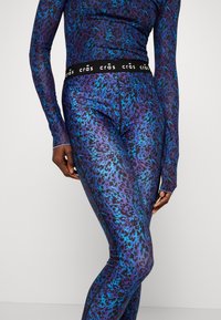 En person i en långärmad, åtsittande outfit med lila och blå djurtryck står mot en vit bakgrund och visar upp designen och passformen.