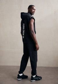 Zwarte mouwloze hoodie met "PUMA CREW '48" in het wit op de achterkant, gecombineerd met zwarte broek en zwarte sportschoenen met witte accenten.