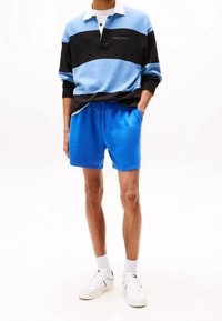 Svetr s dlouhými rukávy, barevně rozlišený v světle modré, tmavě modré a černé barvě s bílým límcem, doplněný modrými šortkami a bílými teniskami.