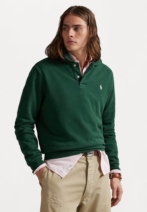 Green long-sleeve pullover with a buttoned collar, featuring a small white logo, worn over a pink checkered shirt and beige pants.