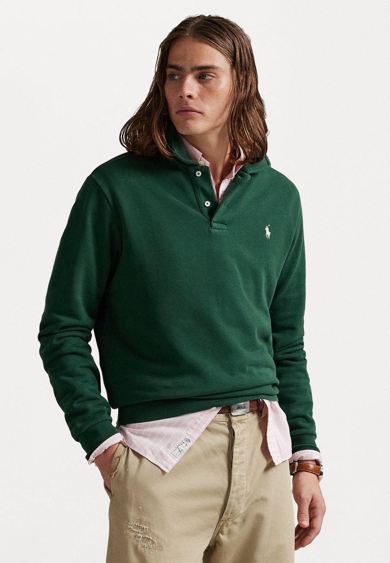 Pull vert à manches longues avec un col à boutons, orné d'un petit logo blanc, porté sur une chemise à carreaux rose et un pantalon beige.