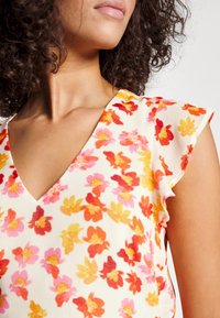 Donna con capelli ricci che indossa un top senza maniche bianco con un motivo floreale rosa, arancione e giallo, rivolta leggermente verso destra.