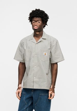 Koszula na guziki z krótkim rękawem w szaro-białe paski, z dwoma kieszeniami na piersi i naszywką z logo. Noszona z niebieskimi spodniami dżinsowymi.