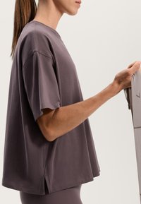 Kvinna i mauve oversized t-shirt som håller i ett ljusgrått motståndsband, visad i sidoprofil med håret uppsatt i hästsvans.