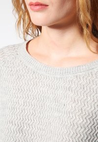 Ljusgrå stickad tröja med ett vågigt mönster, rund halsringning och ribbade ärmslut. Mjuk textur och ventilerande design.