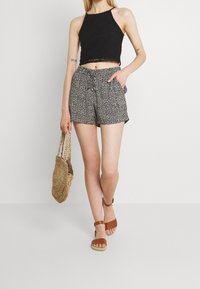 Žena v černém bezramínkovém crop topu, vzorovaných šortkách s vysokým pasem, hnědých sandálech, držící tkanou slaměnou tašku, stojí na pozadí s jednobarevným pozadím.