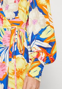 Vestido de estampado floral con colores vibrantes como azul, naranja y rosa. Tela texturizada con un lazo en la cintura y mangas recogidas.