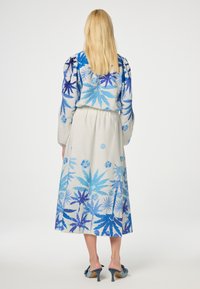 Lang jurk met een blauw bloemenpatroon op een lichte achtergrond, met pofmouwen en een verzamelde taille. Draagt het met gestructureerde blauwe hakken.