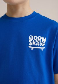 Blå bomullströja med korta ärmar, med en vit grafisk design som säger "BORN TO SKATE" ovanför en skateboardillustration.