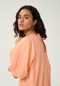 Perzikkleurig, loszittend topje met korte mouwen en een striksluiting op de rug. Zachte textuur en lichte drape in het ontwerp.