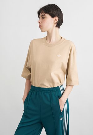 Nuori henkilö, jolla on lyhyet tummat hiukset, beige Adidas t-paita ja petroolinväriset Adidas verkkarit valkoisilla raidoilla, kädet taskuissa, katsoo vasemmalle.