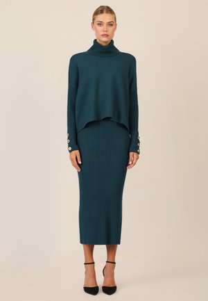 Türkisfarbener gerippter Strick-Turtleneck-Pullover mit überschnittenen Schultern, kombiniert mit einem passgenauen, passenden Rock. Mit Knopfakzenten an den Ärmeln.
