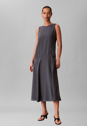 Kobieta stoi, ubrana w bezrękawnikową ciemnoszarą sukienkę midi i czarne sandały na obcasie na jednolitym jasnoszarym tle.