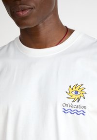 T-shirt de algodão branca com um design colorido de sol com um olho, texto "Em Férias", e gráficos de ondas azuis no lado esquerdo.