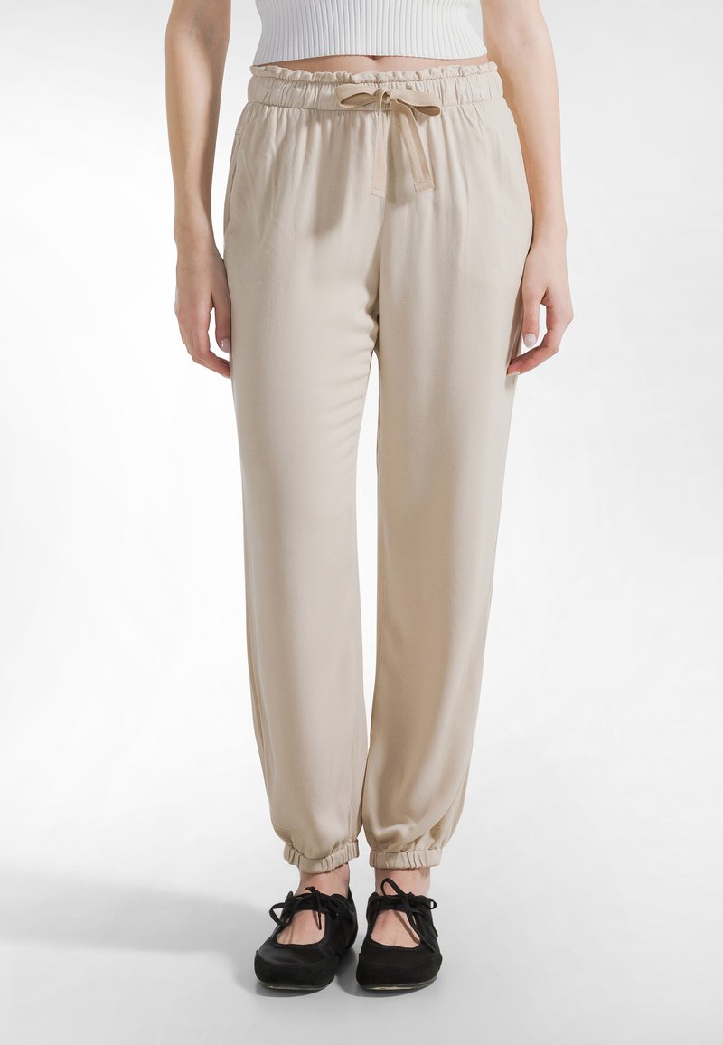 Beige, lette joggingbukser med elastisk linning og snøre. Har elastiske manchetter ved anklerne. Fremstillet af glat stof.