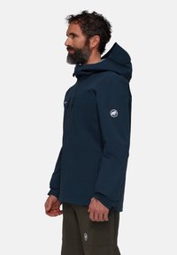 Veste imperméable bleu marine avec capuche, dotée d'une fermeture éclair à l'avant, d'une poche sur la poitrine et d'un patch logo sur la manche. Texture lisse et design minimaliste.