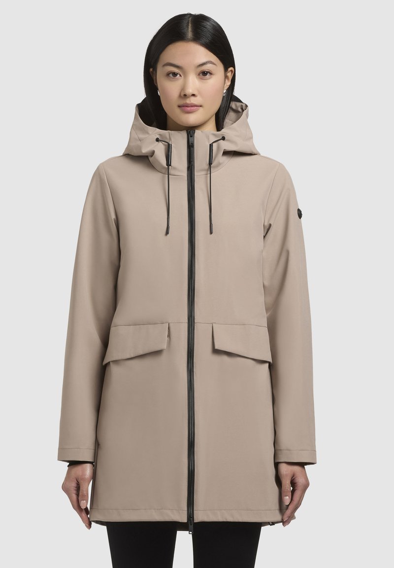 Beige vandtæt jakke med hætte, frontlynlås, to store lommer og kontrasterende sorte metaldele. Glat tekstur og ustruktureret design.