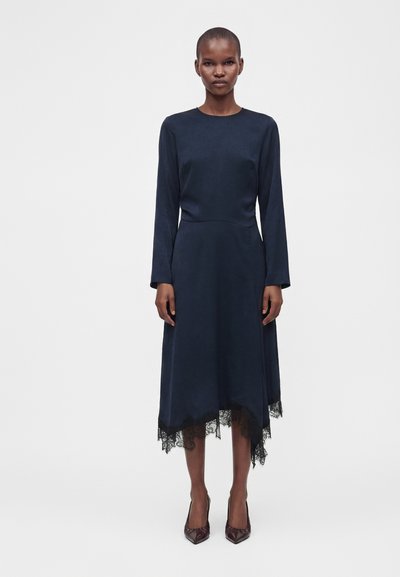 Femme portant une robe bleu marine à manches longues avec un ourlet en dentelle asymétrique et des talons hauts pointus marron foncé, se tenant devant un fond uni.