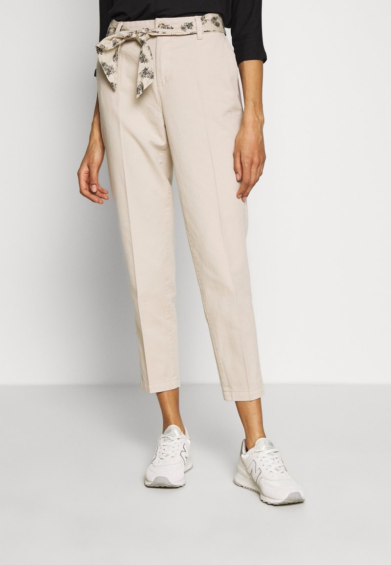 Pantalon beige court avec une ceinture en tissu à motif floral nouée à la taille, associé à des baskets blanches et un haut noir.