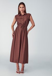 Brown, sleeveless maxi dress with button-front, two chest pockets, gathered waist, and white polka dots. Paired with white heeled sandals.