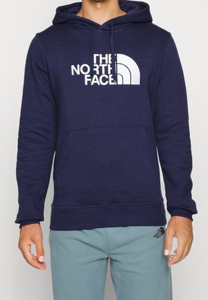 Homme portant un sweat à capuche bleu marine The North Face avec poche avant et pantalon bleu clair avec un petit logo noir sur la cuisse gauche.