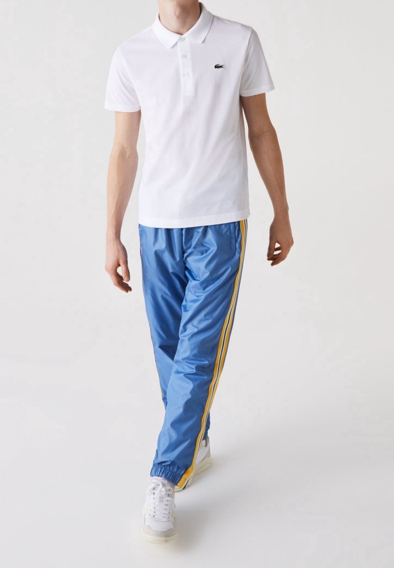 Homme portant un polo blanc à manches courtes et un pantalon de sport bleu avec des bandes jaunes marchant devant un fond blanc uni.