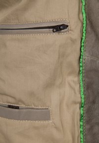 Tessuto del giubbotto beige con una toppa in pelle marrone, dotato di due tasche con zip e cuciture verdi lungo la cucitura per un contrasto.
