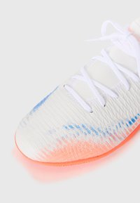 Embout d'une chaussure de sport blanche avec surface texturée, lacets blancs, motif dégradé bleu et orange, et semelle orange visible sur fond blanc.