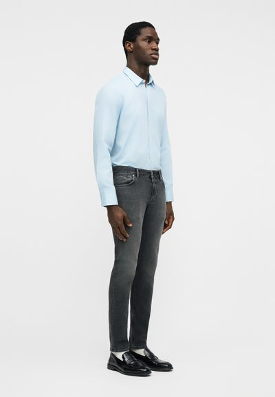 Άνδρας που στέκεται και κοιτά προς τα δεξιά, φορώντας ανοιχτό γαλάζιο πουκάμισο με μακριά μανίκια, σκούρα γκρι τζιν, λευκές κάλτσες και μαύρα μοκασίνια, με φόντο απλό.