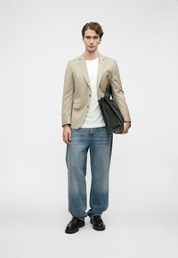 Νεαρός άνδρας που φοράει μπεζ σακάκι, λευκό μπλουζάκι, φαρδιά μπλε τζην και μαύρα παπούτσια, κρατώντας μια μεγάλη μαύρη δερμάτινη τσάντα.