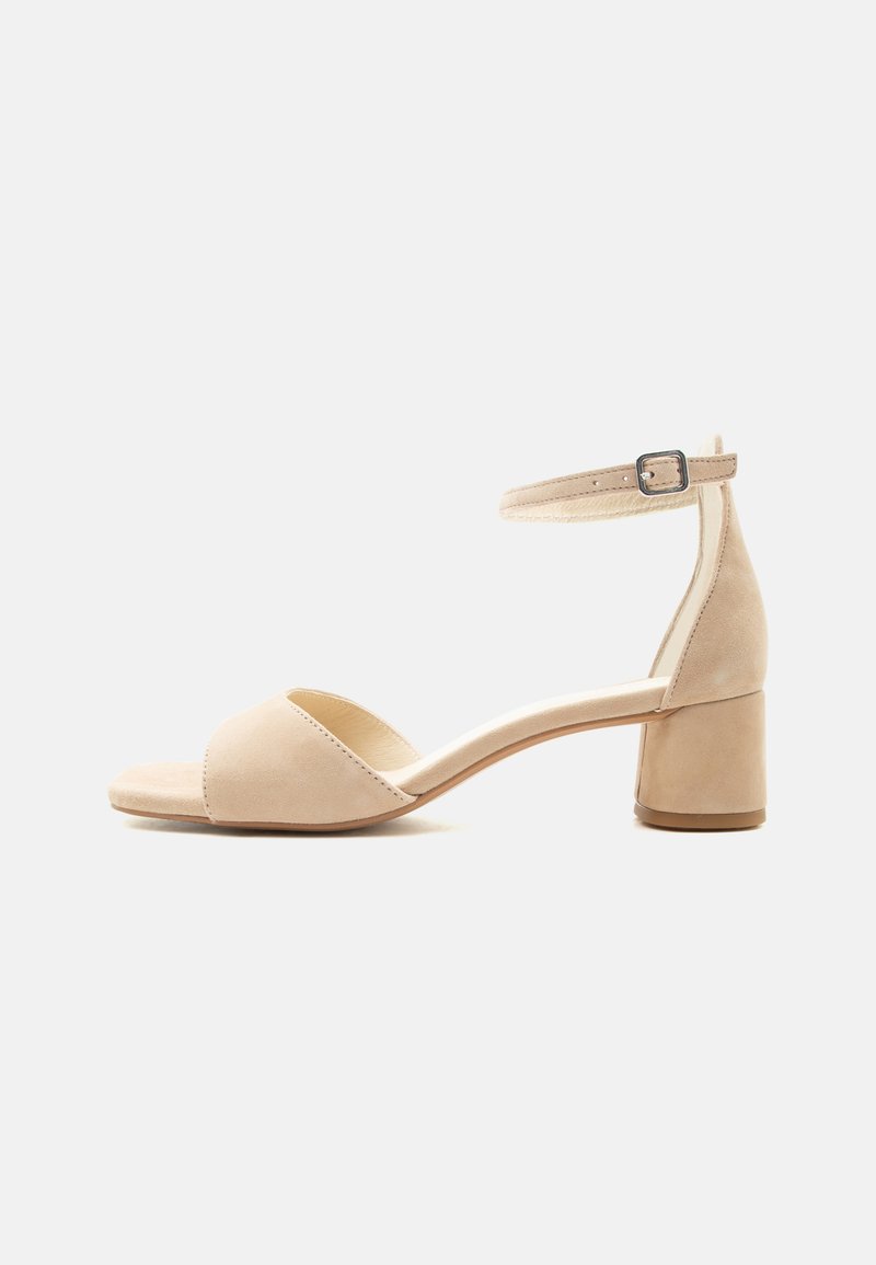 Μπεζ σουέντ ανοιχτή σανδάλι με χοντρό τακούνι και λουράκι στον αστράγαλο με μικρή μεταλλική αγκράφα, εμφανίζεται σε προφίλ σε λευκό φόντο.