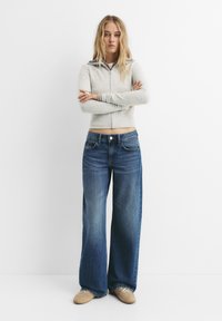 Svetlo sivi zip-up pulover z rebrasto teksturo, v kombinaciji s širokimi modrimi denim hlačami. Hlače imajo gumbno zapiranje in sprednje žepke.