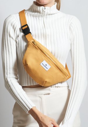 Gul lærred sling taske med lynlåslukning og en hvid logo-patch, der har en tekstureret overflade og en justerbar rem.