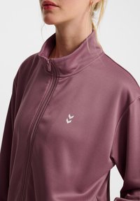 Lilla zip-up jakke lavet af glat stof, med høj krave og et diskret logo. Nærbillede viser lynlåsdetalje og tekstur.