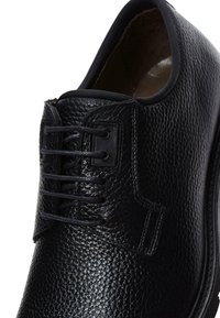 Zapato de cuero negro con un acabado texturizado, que presenta seis ojales, cordones negros y una puntera redondeada. Forro interior suave.