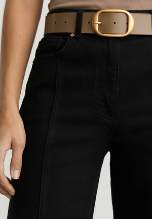 Persoană purtând blugi negri cu talie înaltă și o curea maro cu o cataramă mare, rotundă, aurie, mâna odihnindu-se lângă blugi.