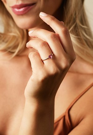 Goldring mit einem herzförmigen rosa Edelstein, der am Ringfinger liegt. Glatte Textur mit minimalistischem Design, vor einem neutralen Hintergrund.