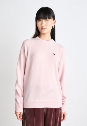 Lichtroze trui met lange mouwen, ronde hals, geribde boorden en zoom. Bevat een klein groen Lacoste-logo op de linkerkant van de borst. Zachte textuur.