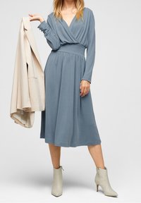 Kobieta w niebieskiej sukience midi i beżowych botkach do kostek trzyma złożony beżowy płaszcz na jednym ramieniu na tle jednolitym.