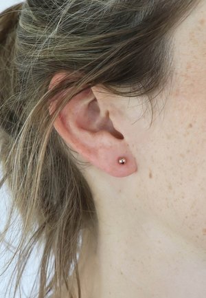 Rosegold Ohrstecker im Ohrläppchen platziert; sphärische Form, glatte Textur, kleine Größe, minimalistisches Design, das den Hautton und das Haar ergänzt.