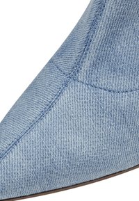 Gros plan d'une chaussure en denim bleu clair à bout pointu montrant des coutures détaillées et la texture du tissu sur la partie supérieure de la chaussure.