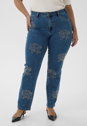 Kvinde iført blå jeans med sølvrose-mønstre kombineret med sorte spidse hæle og en hvid top, stående med den ene hånd på hoften.