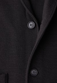 Svart texturerad tygblazer med notchkrage, en knappstängning och en frontficka. Knappen har fyra hål och ingraverade detaljer.